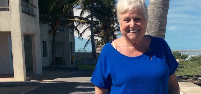 Photo of Helga, an older adult woman standing in front of a building with palm trees in the background