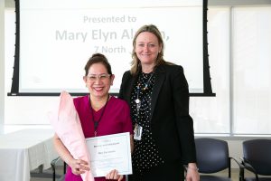 a photo taken at an awards ceremony showing a nurse wearing scrubs holding a bouquet of flowers and a certificate and standing with the Chief Nursing Executive