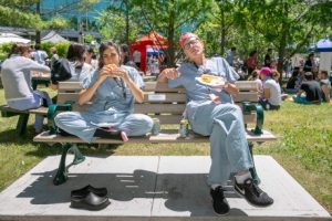 Two people wearing surgical scrubs sit relaxing on a bench outside in a park eating hamburgers 