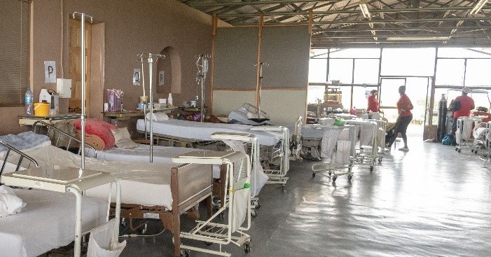 Image shows large open room with hospital beds and equipment along one side. There are three people standing in the background at the back of the room in front of a large window