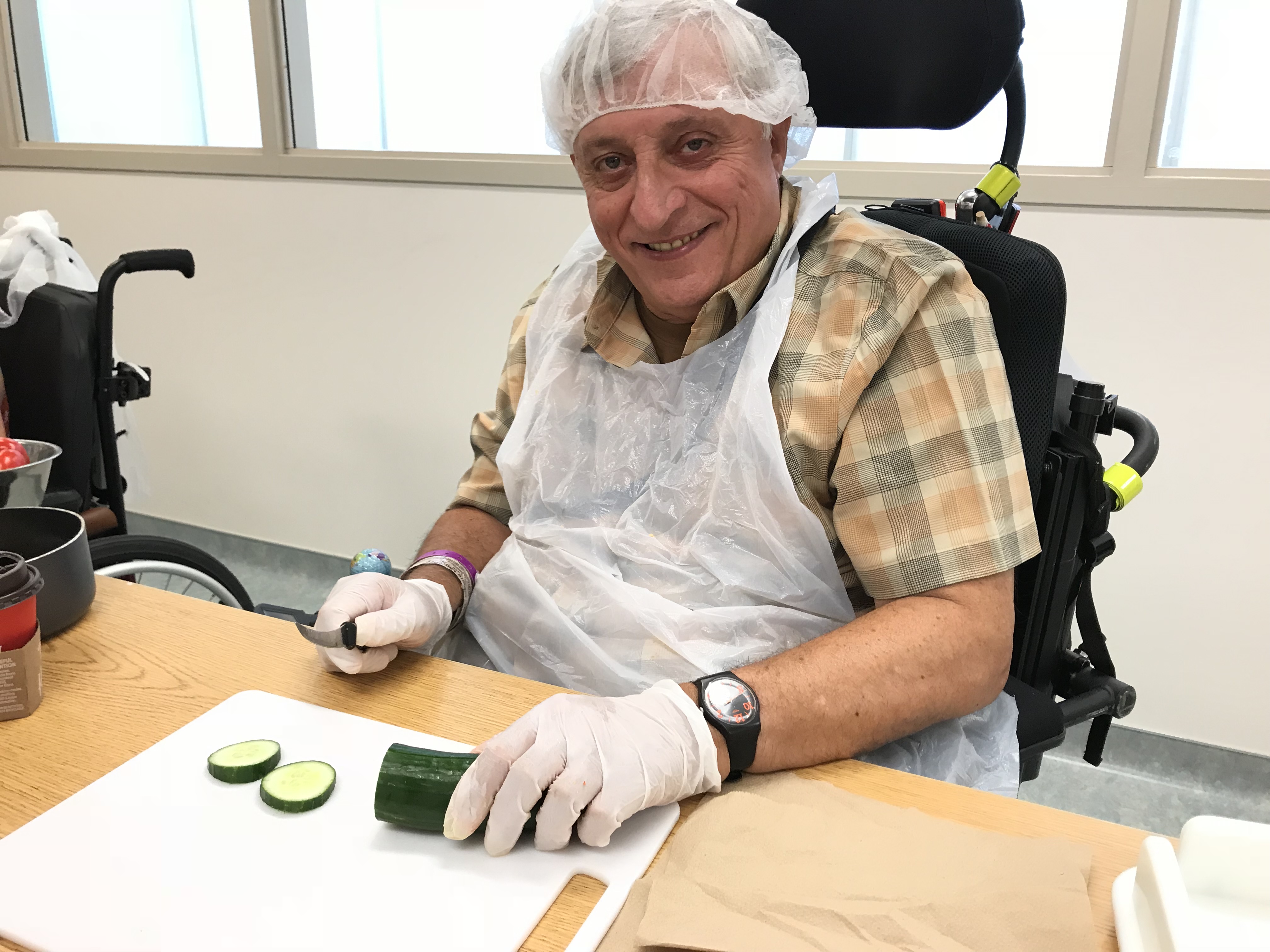A man wearing an apron, hair net and gloves chops a cucumber at a table. 