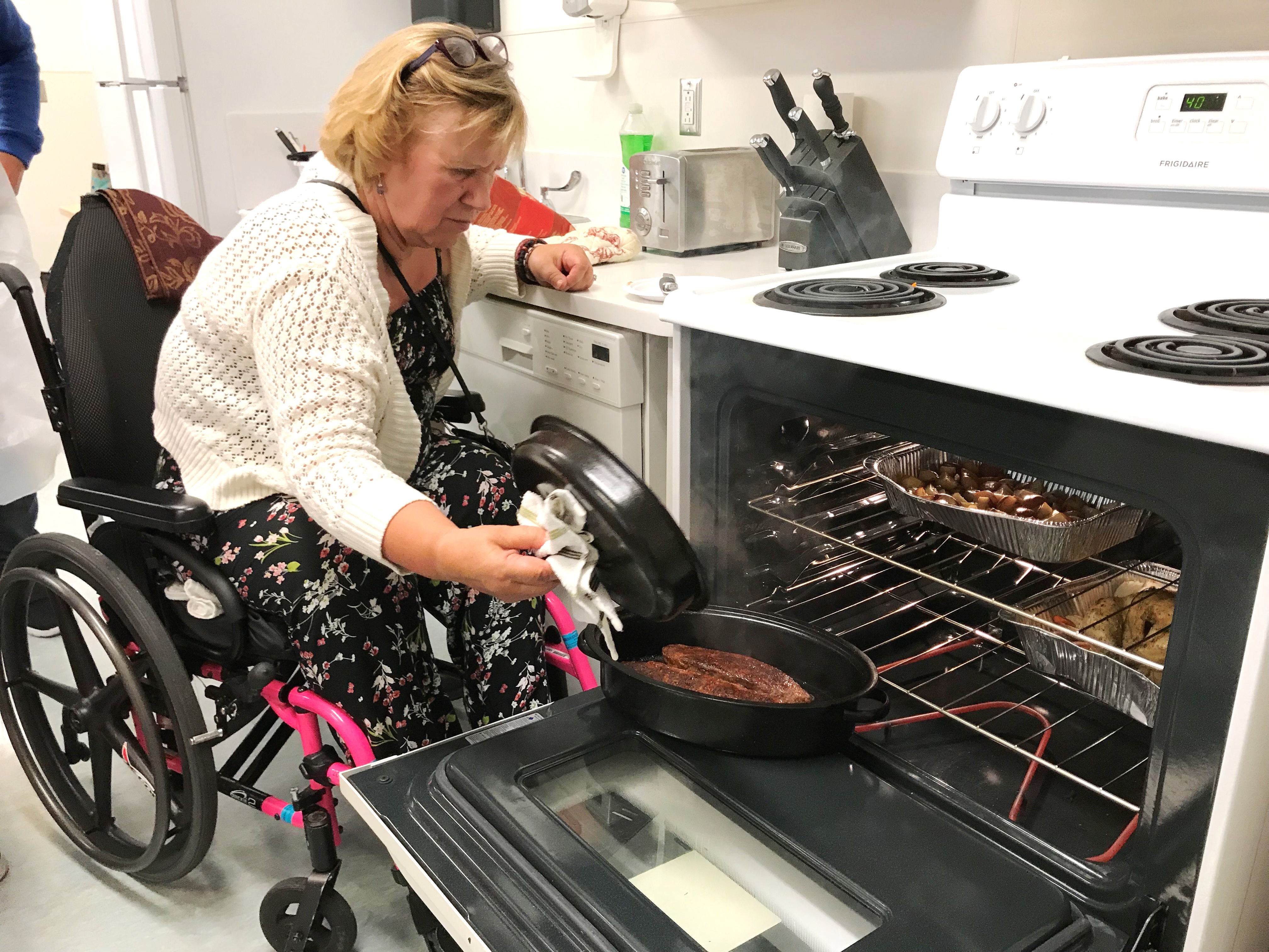 a woman in a wheelchair is next to an open oven. She is taking the lid off a pot to check on the food that's cooking