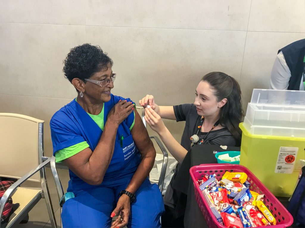 An employee wearing hospital scrubs sits next to a table that has mini chocolate bars and a yellow plastic container on it. She's holding her sleeve rolled up and looking at another employee who is next to her putting a cotton ball on her arm after giving her the flu shot.